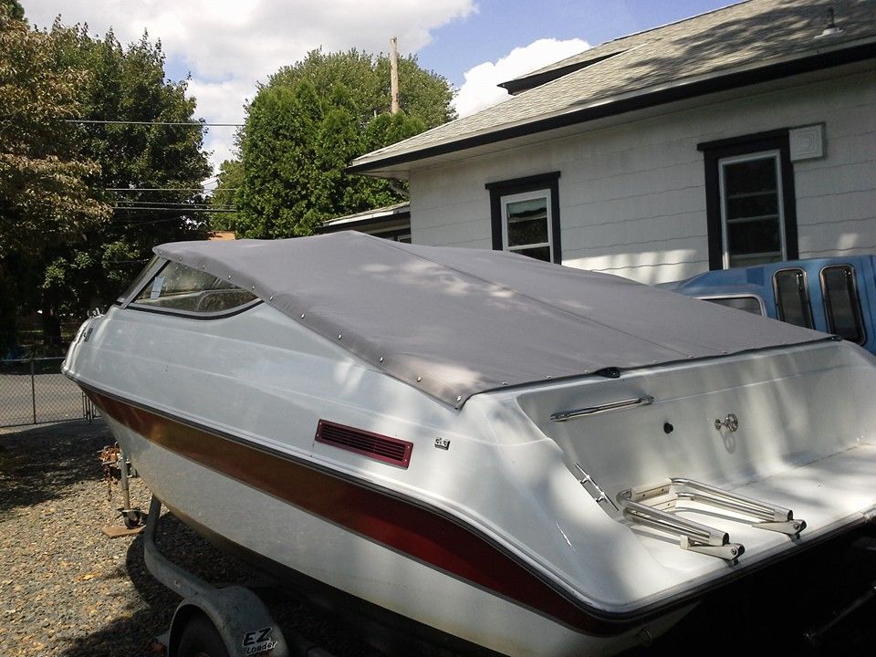 Silver boat on a trailer in a driveway beside a house, with a red stripe and covered bow.