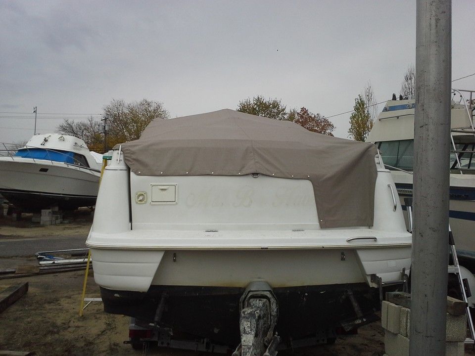 White boat with beige cover on trailer in a storage yard, with another boat and a utility pole nearby.