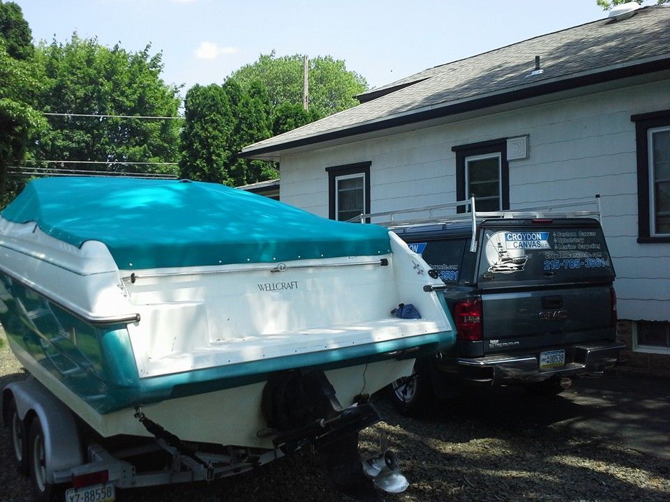 Boat on trailer beside black pickup truck next to a white house, with blue cover and green trees in back.