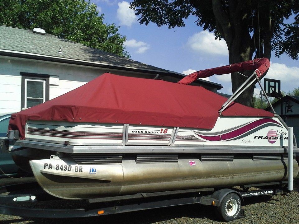 Red pontoon boat on a trailer parked beside a house under trees and blue sky