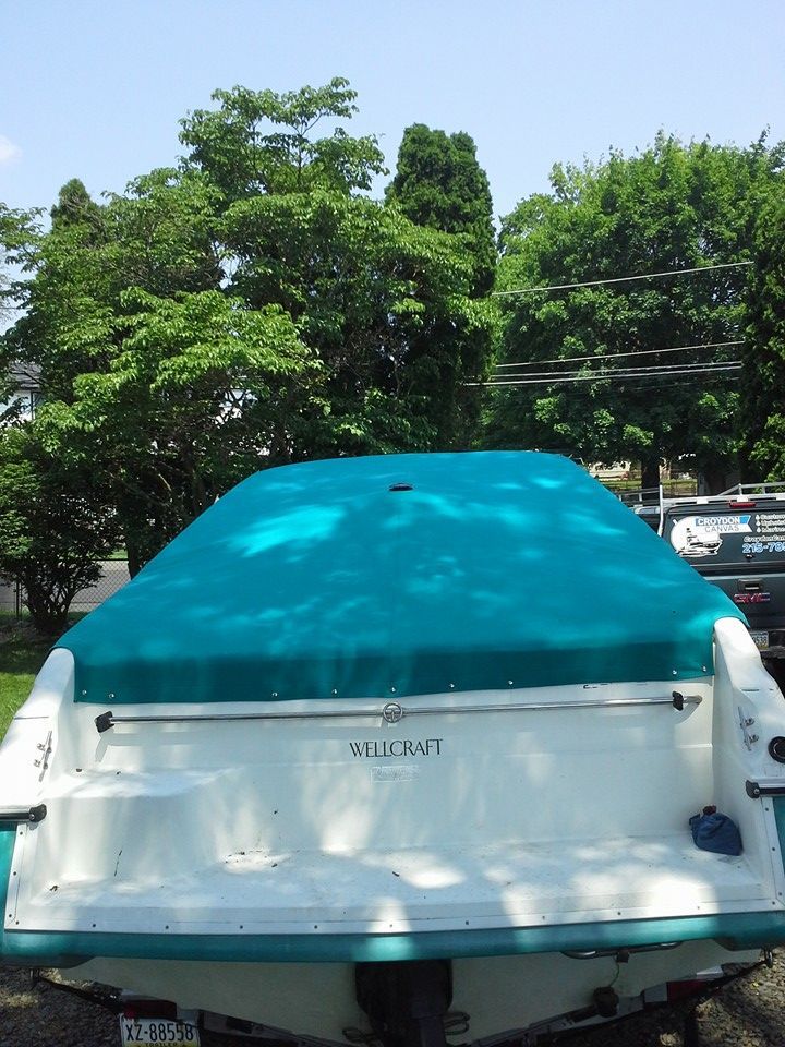 Rear view of a white boat with a teal cover parked outdoors среди trees and parked cars
