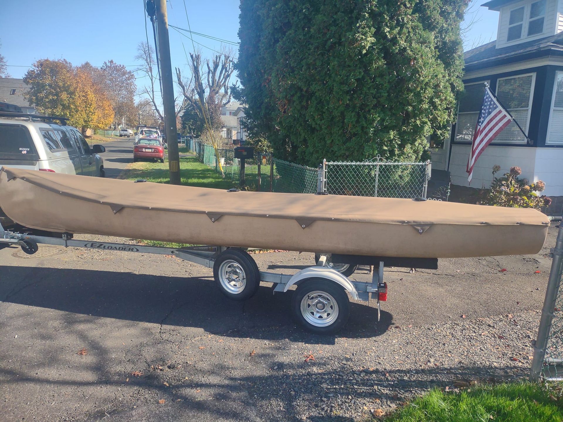 Tan canoe on a small trailer in a gravel driveway beside a house and American flag.