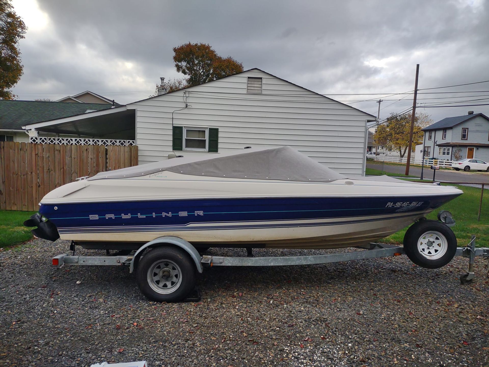 Blue-and-white speedboat on a trailer in a gravel yard, with a house and cloudy sky behind.