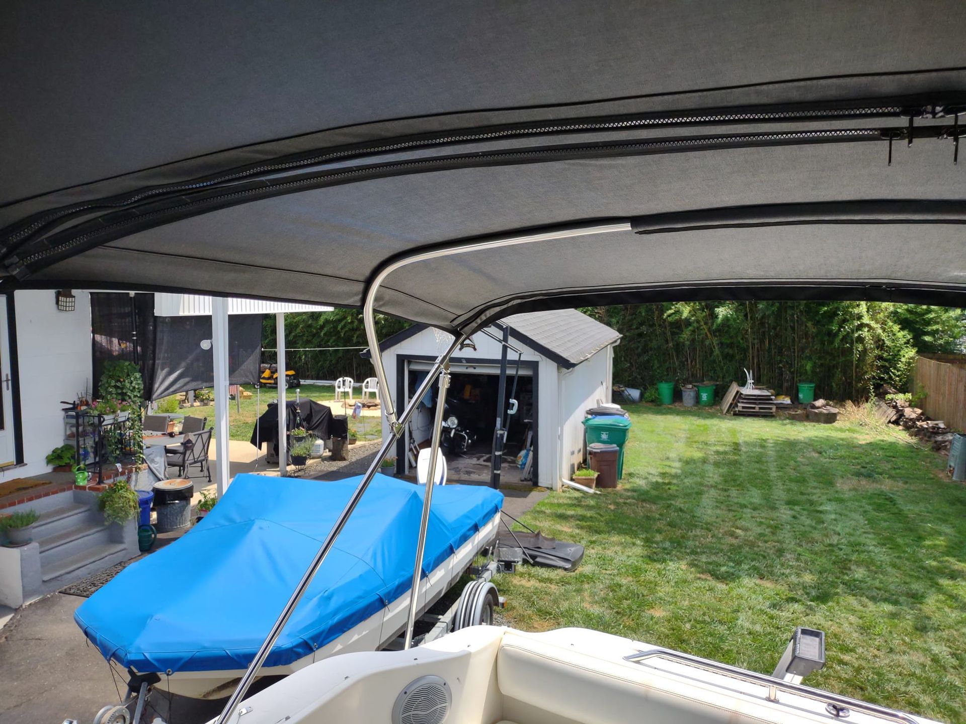 Boat under a carport beside a house, with a blue cover and a grassy yard in the background