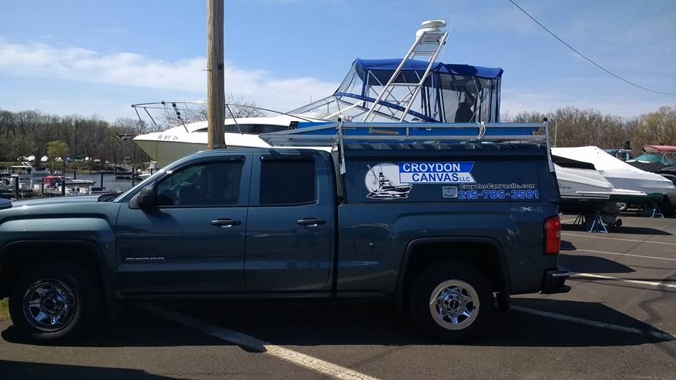 Blue pickup truck with a canopy parked in a lot beside several boats on trailers