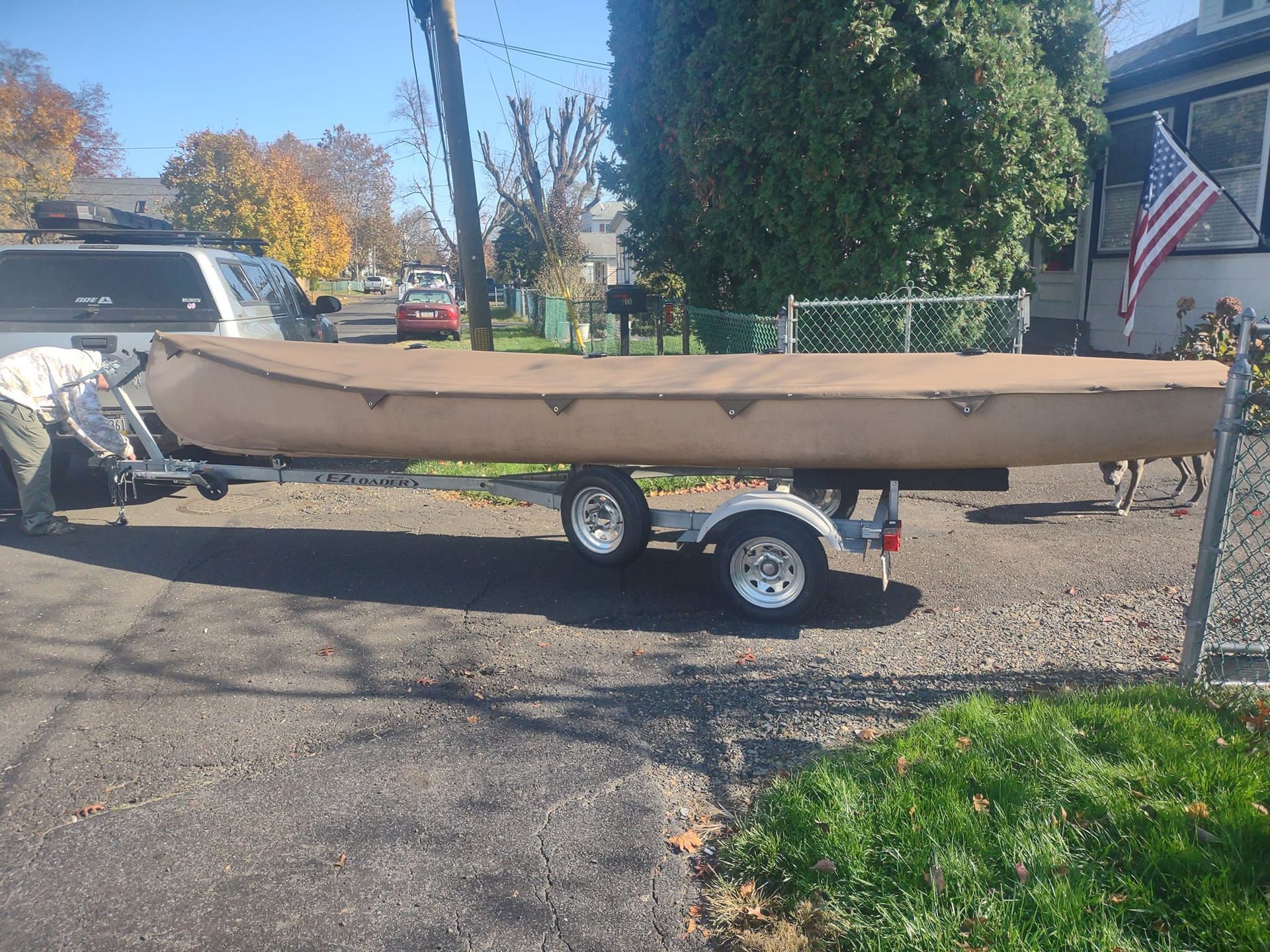 Brown canoe on a small trailer parked in a driveway, with autumn trees and a U.S. flag in the background