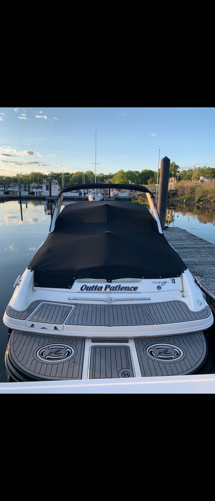 White speedboat with a black cover docked on calm water at sunset, marina and trees in background