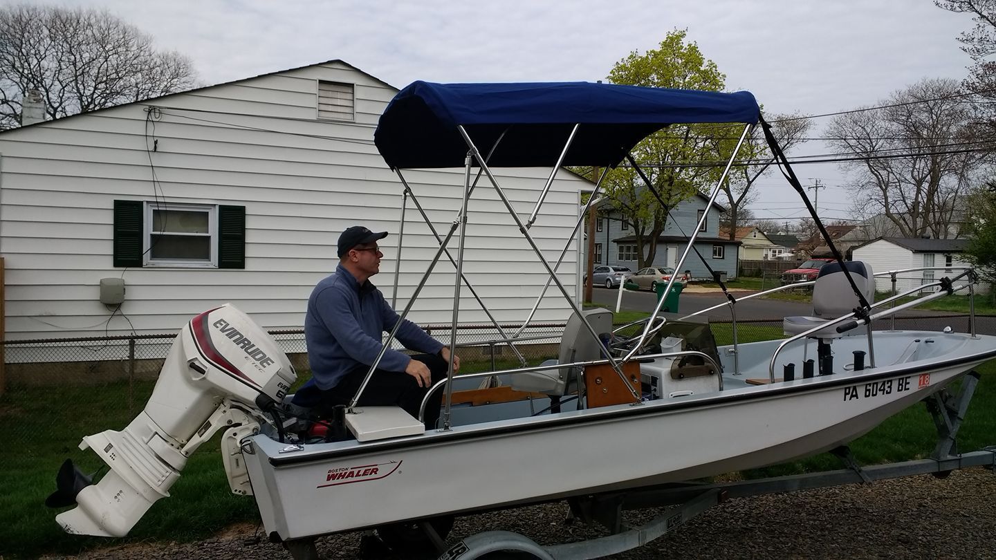 Small motorboat on a trailer in a yard, with a person sitting inside under a blue canopy.