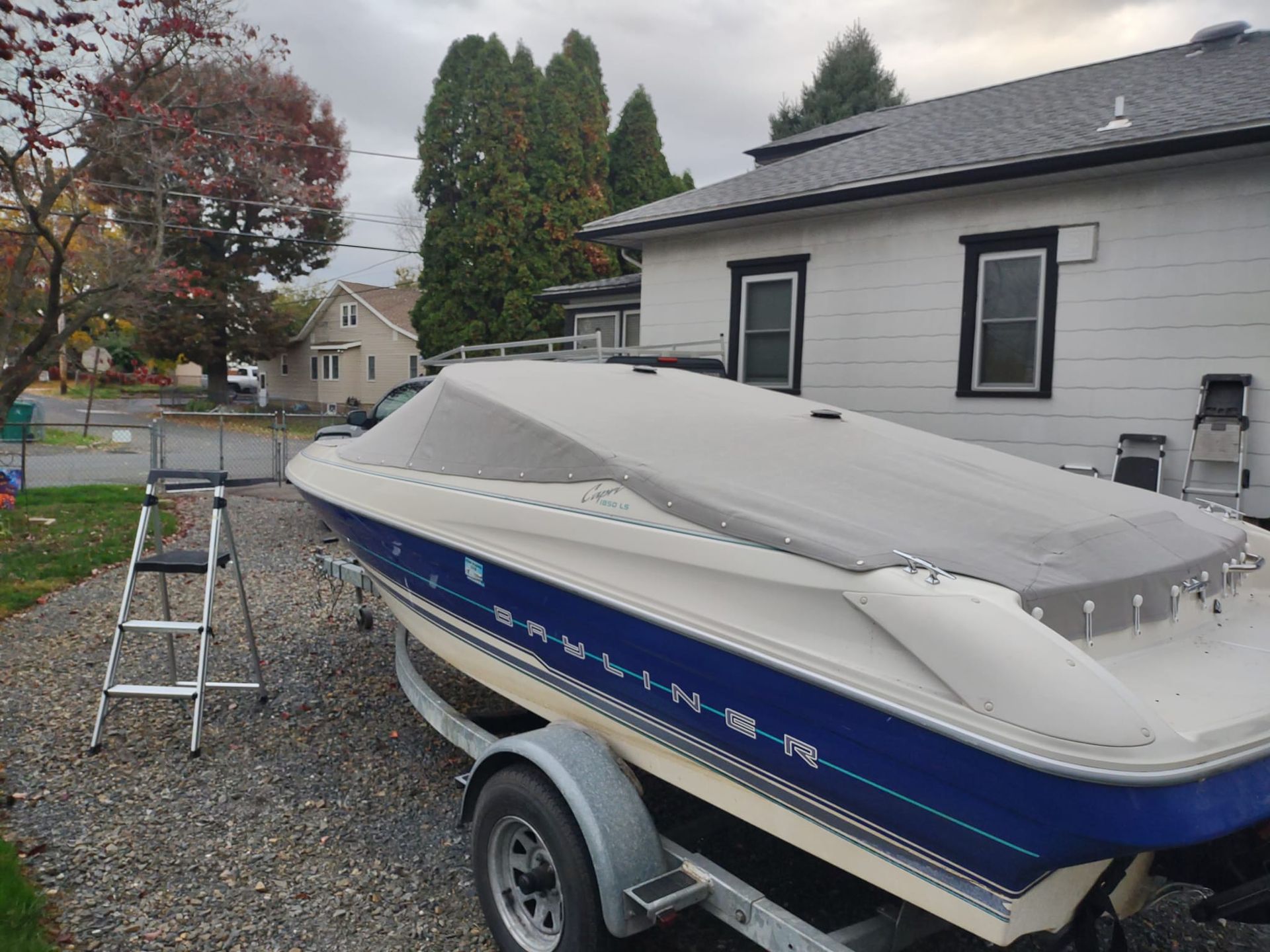 Covered white-and-blue boat on a trailer beside a house, with a ladder in the driveway.