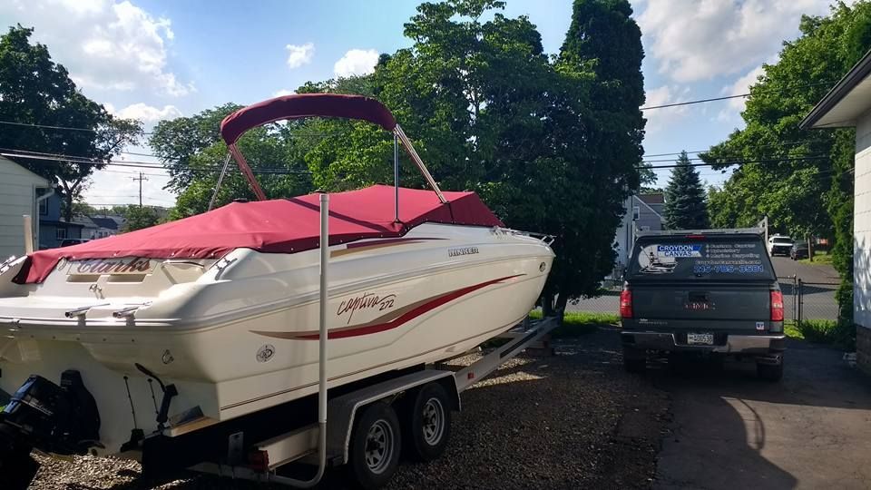 White speedboat on trailer beside a pickup truck in a driveway, with trees and red cover in the background