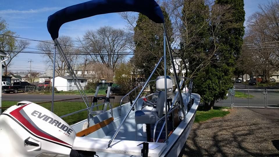Small white sailboat on a trailer with a blue canopy, parked outdoors near trees and houses.