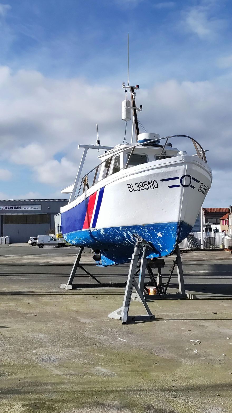 Small white and blue boat on a cradle in a shipyard under a partly cloudy sky
