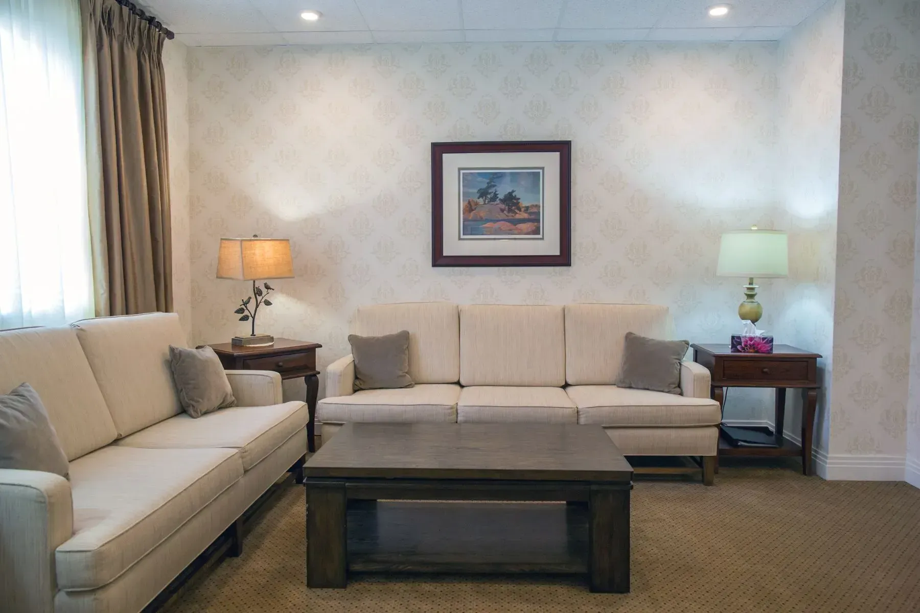 Bright living room with beige sofas, dark coffee table, and lamps on either side of a framed wall print.