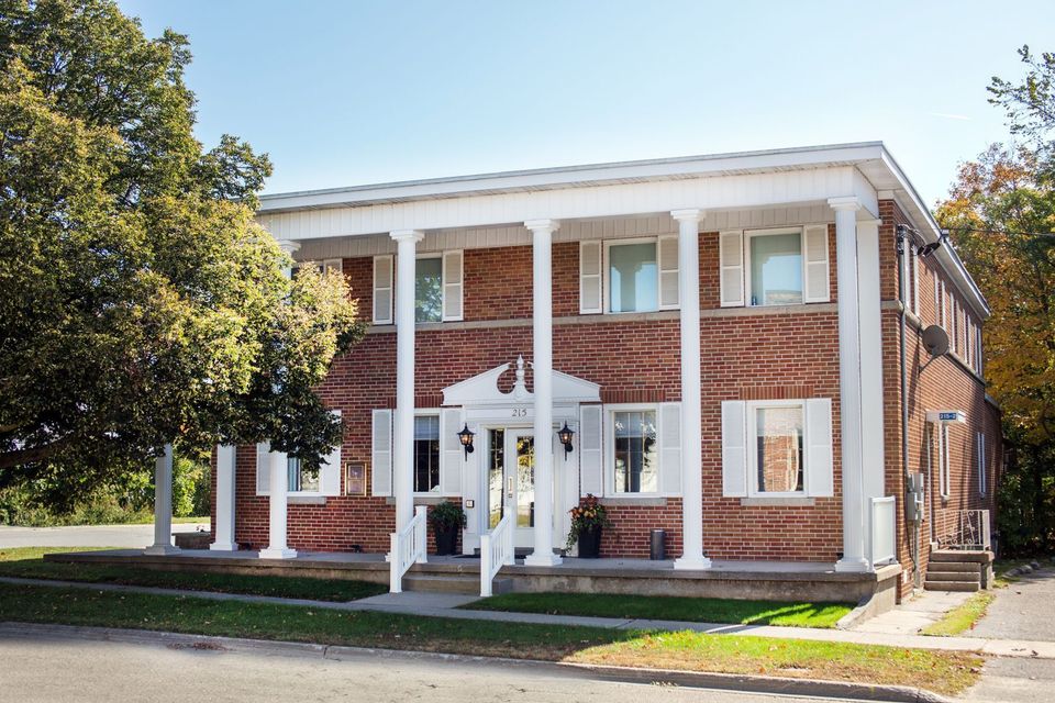 Brick two-story building with white columns, front porch, and trees along the sidewalk.