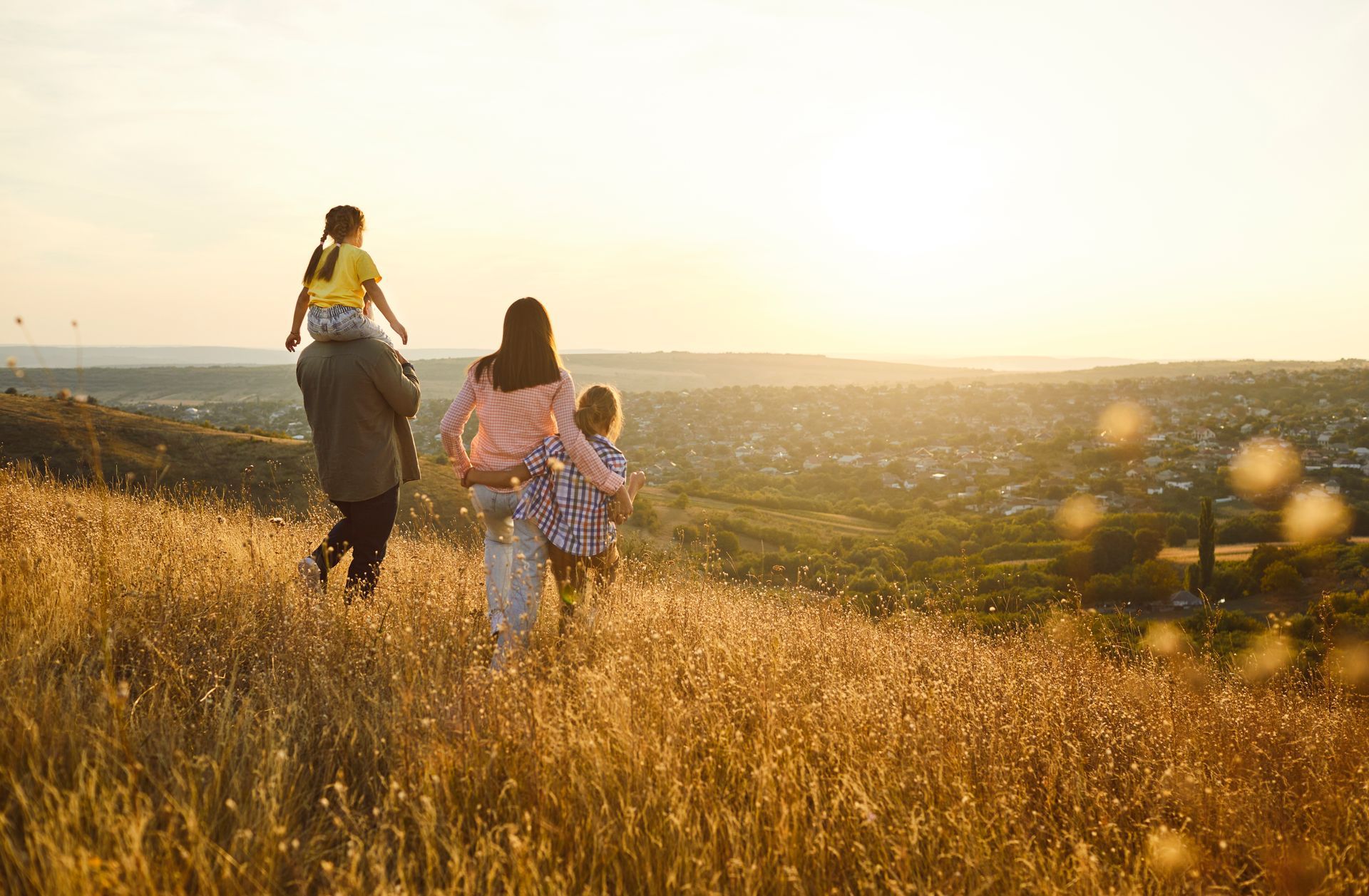 Three people walking through a golden field at sunset, carrying a child on one person’s shoulders.