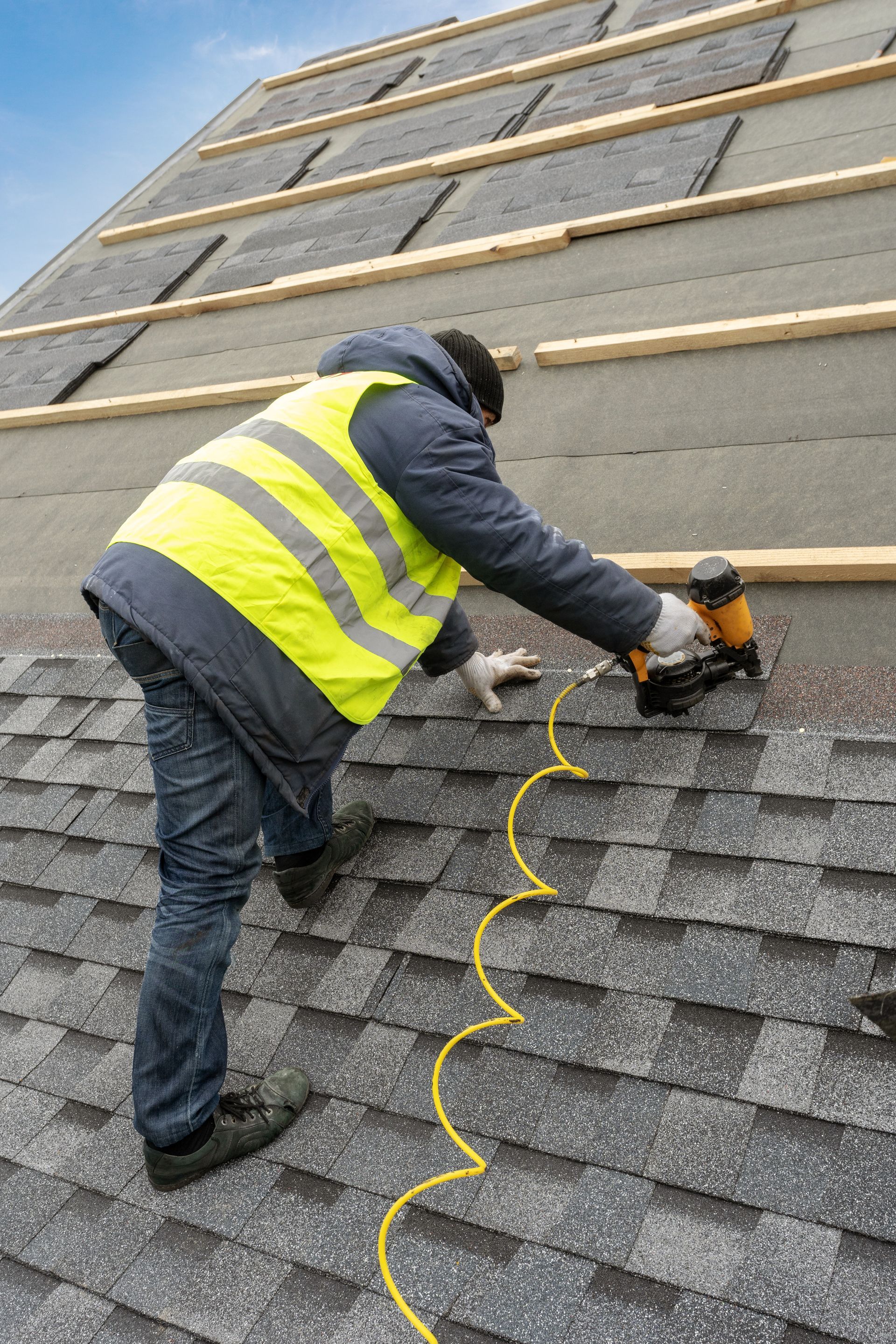 Worker in a yellow safety vest using a nail gun on a shingled roof