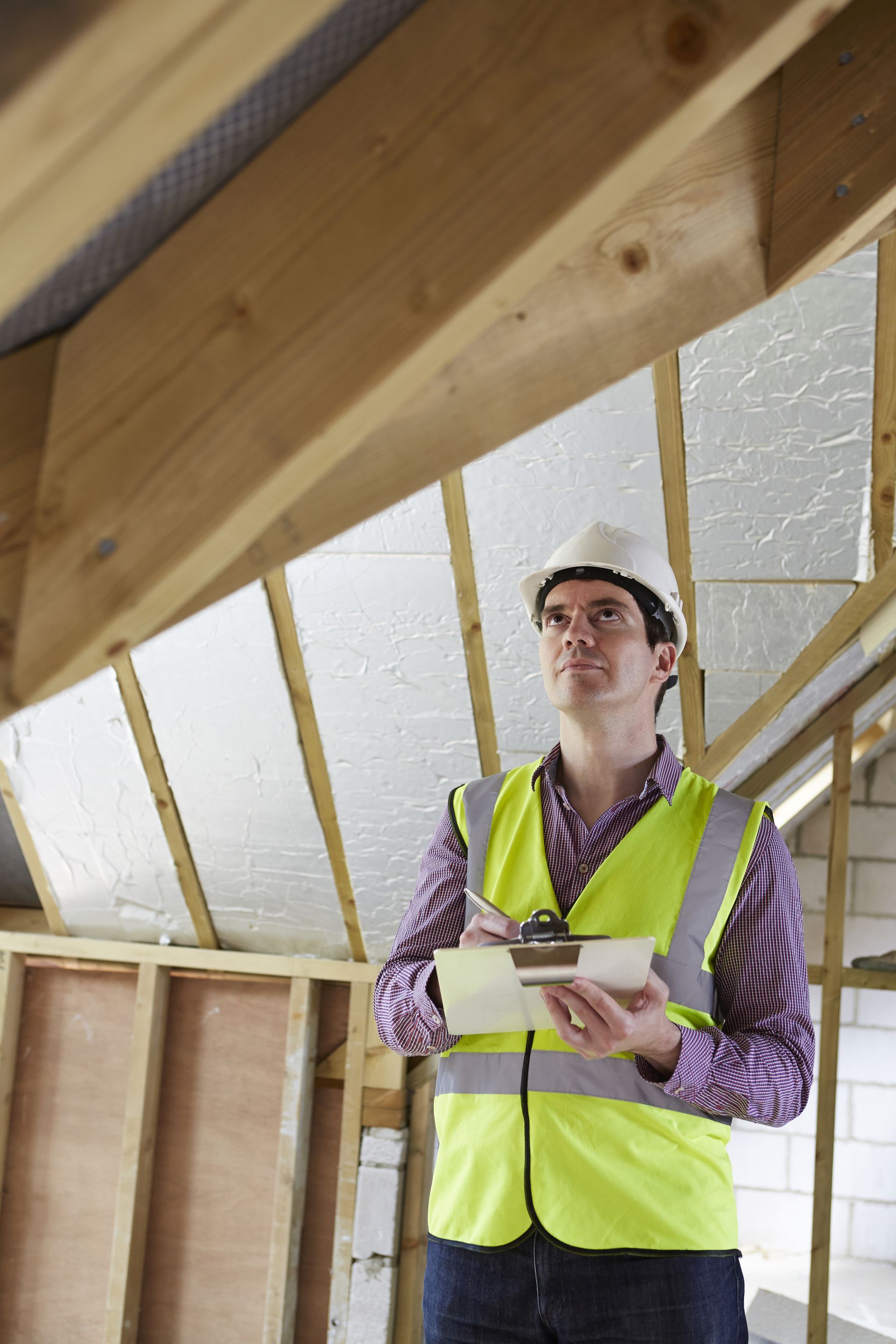 Construction worker in a hard hat and safety vest holding a clipboard inside an unfinished attic room