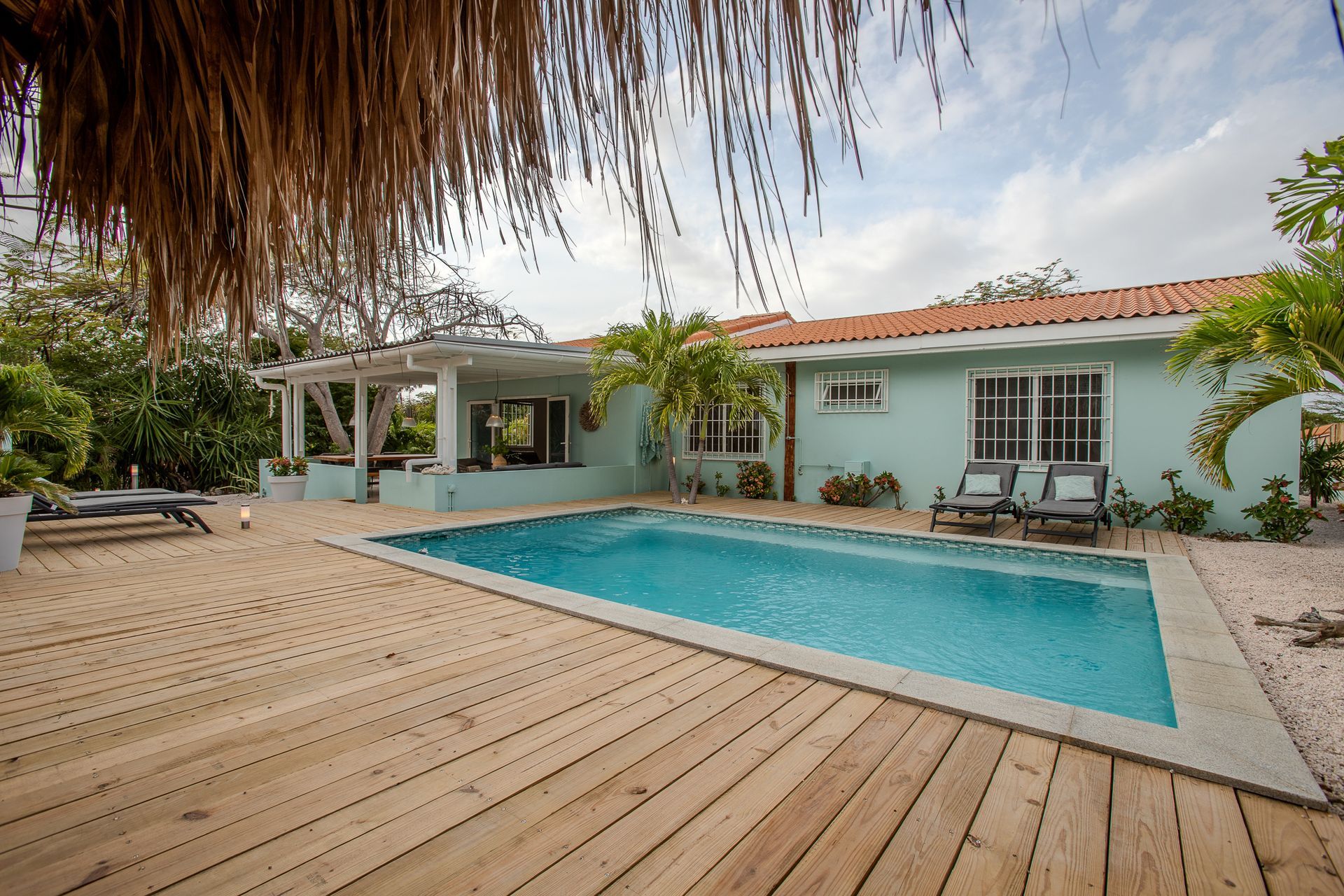 Een turquoise huis met zwembad, houten terras en een rieten dakoverhang.