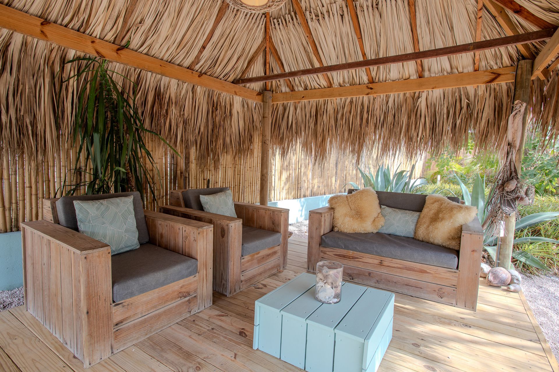 Buiten zitgedeelte met houten meubilair, een rieten dak en een turquoise tafel.