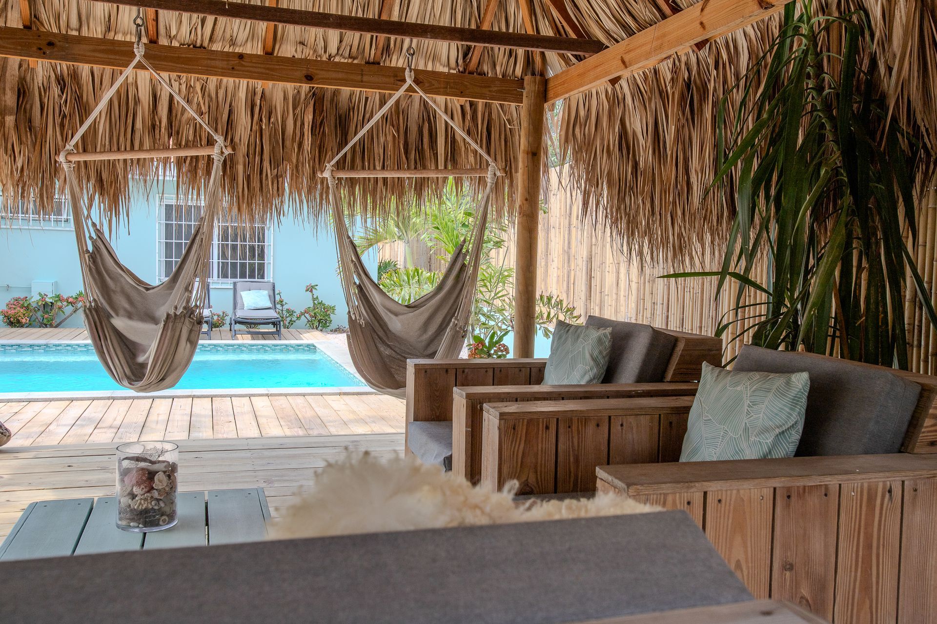 Een loungegedeelte bij het zwembad met houten meubels, hangstoelen, een rieten dak en het zwembad op de achtergrond.