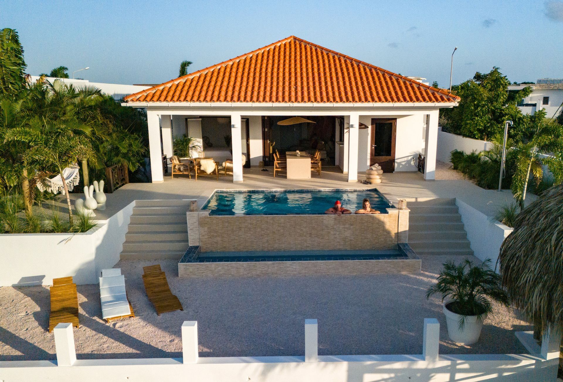 Wit strandhuis met oranje pannendak, zwembad en ligstoelen.