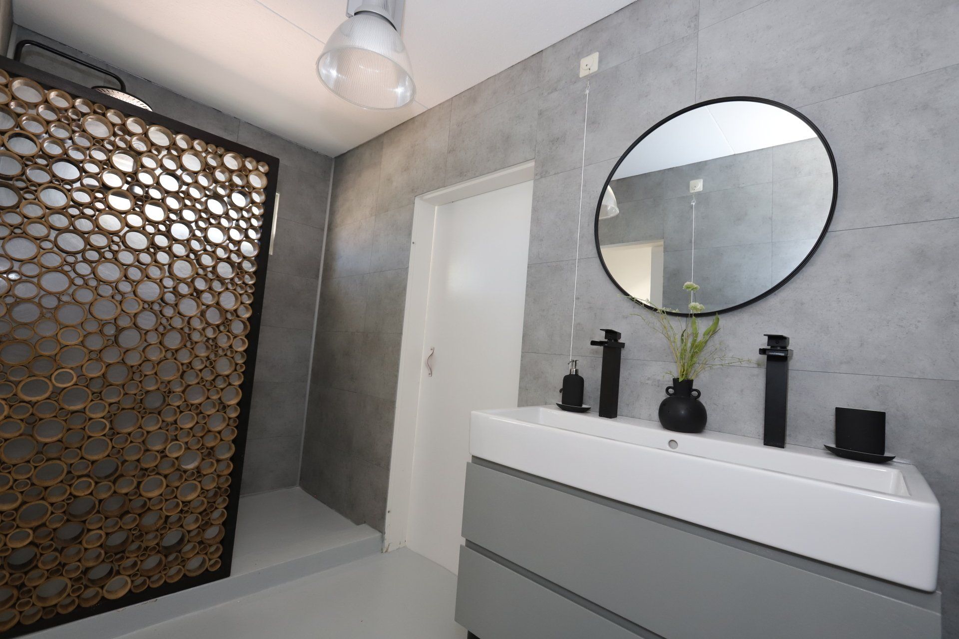 Moderne badkamer met grijze muren, witte wastafel, ronde spiegel en decoratief houten kamerscherm.