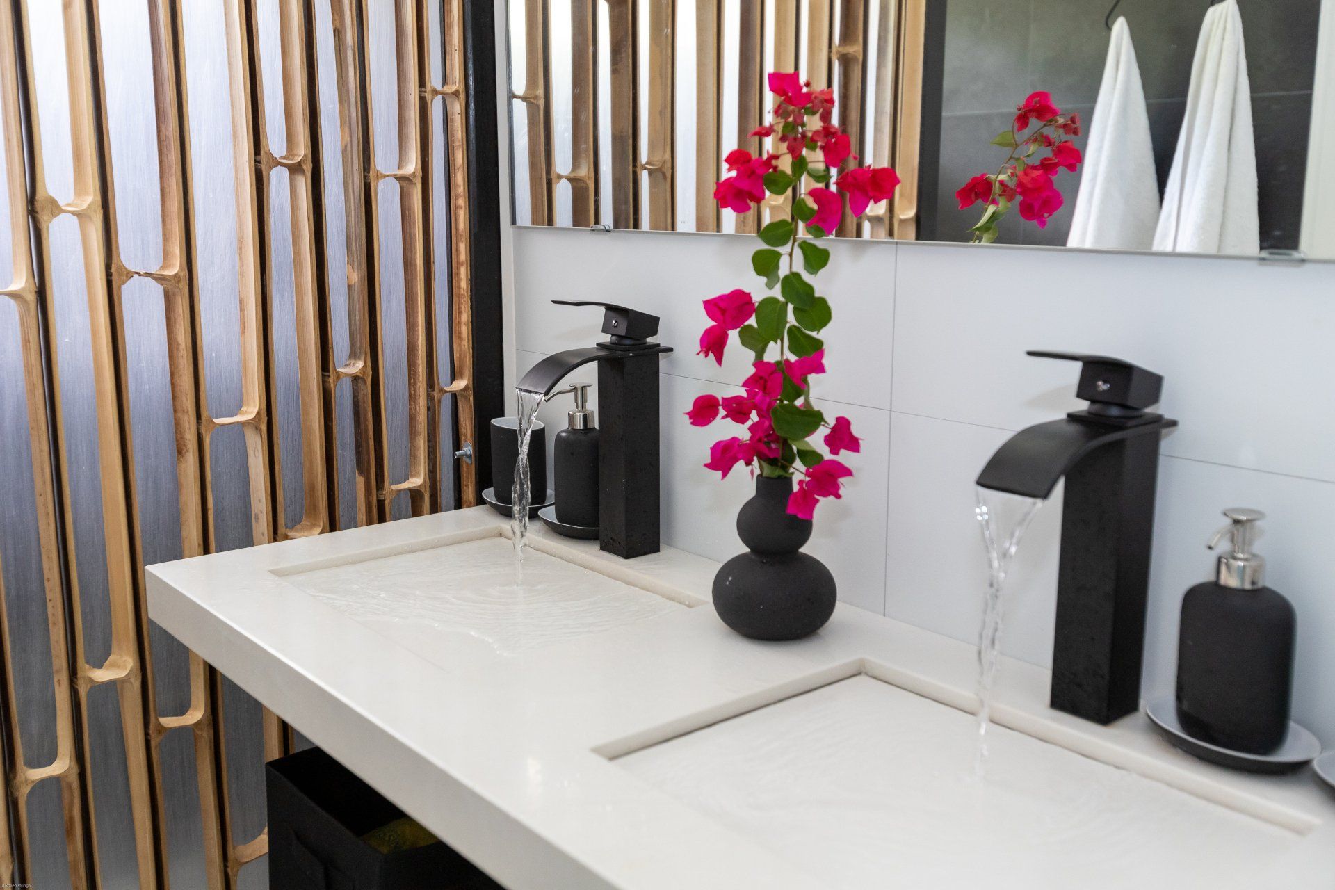 Moderne badkamer met wit aanrechtblad, zwarte kranen, een vaas met roze bloemen en een accentmuur van bamboe.