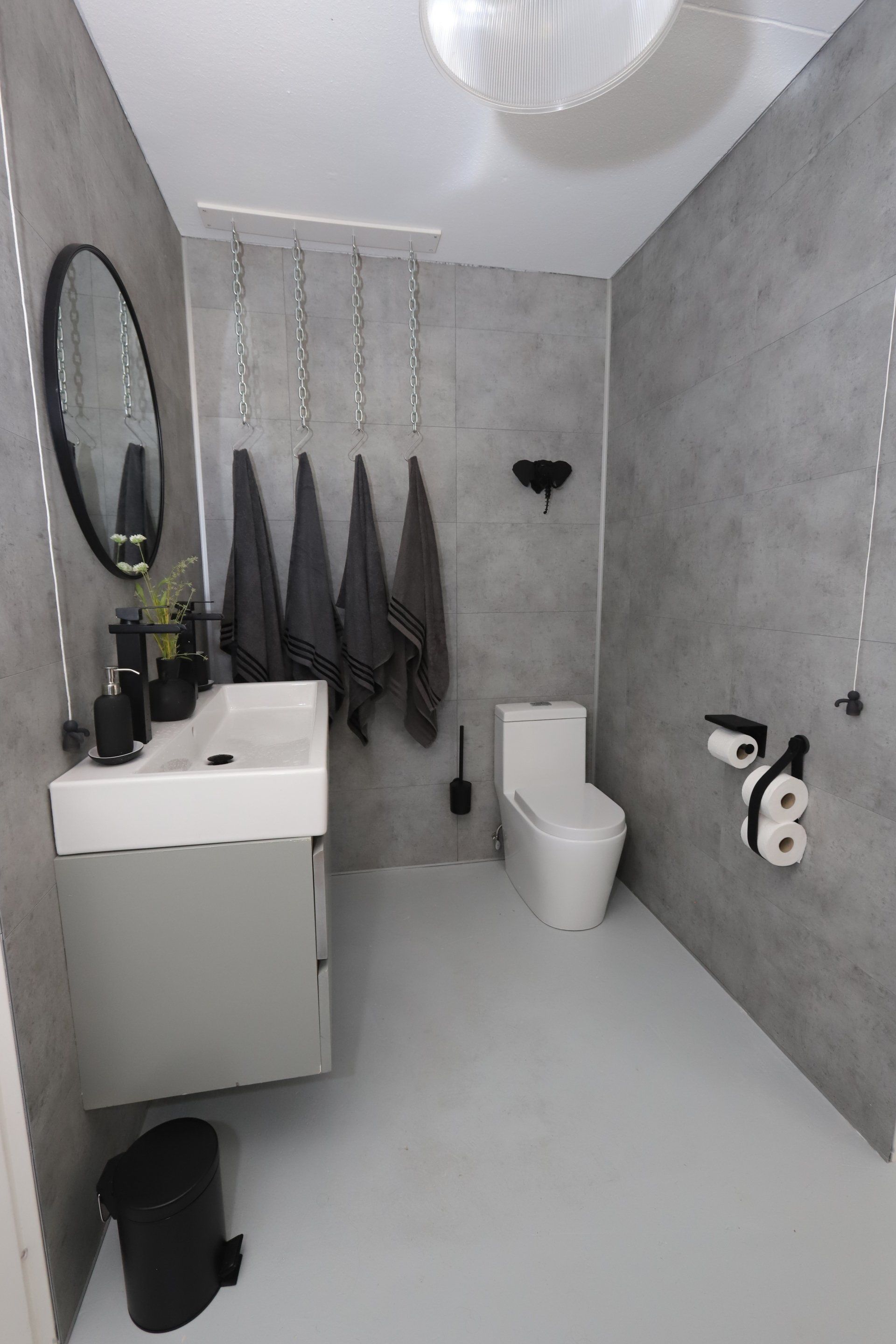 Moderne badkamer met grijze muren, wit toilet en wastafel, zwarte accessoires en hangende handdoeken.