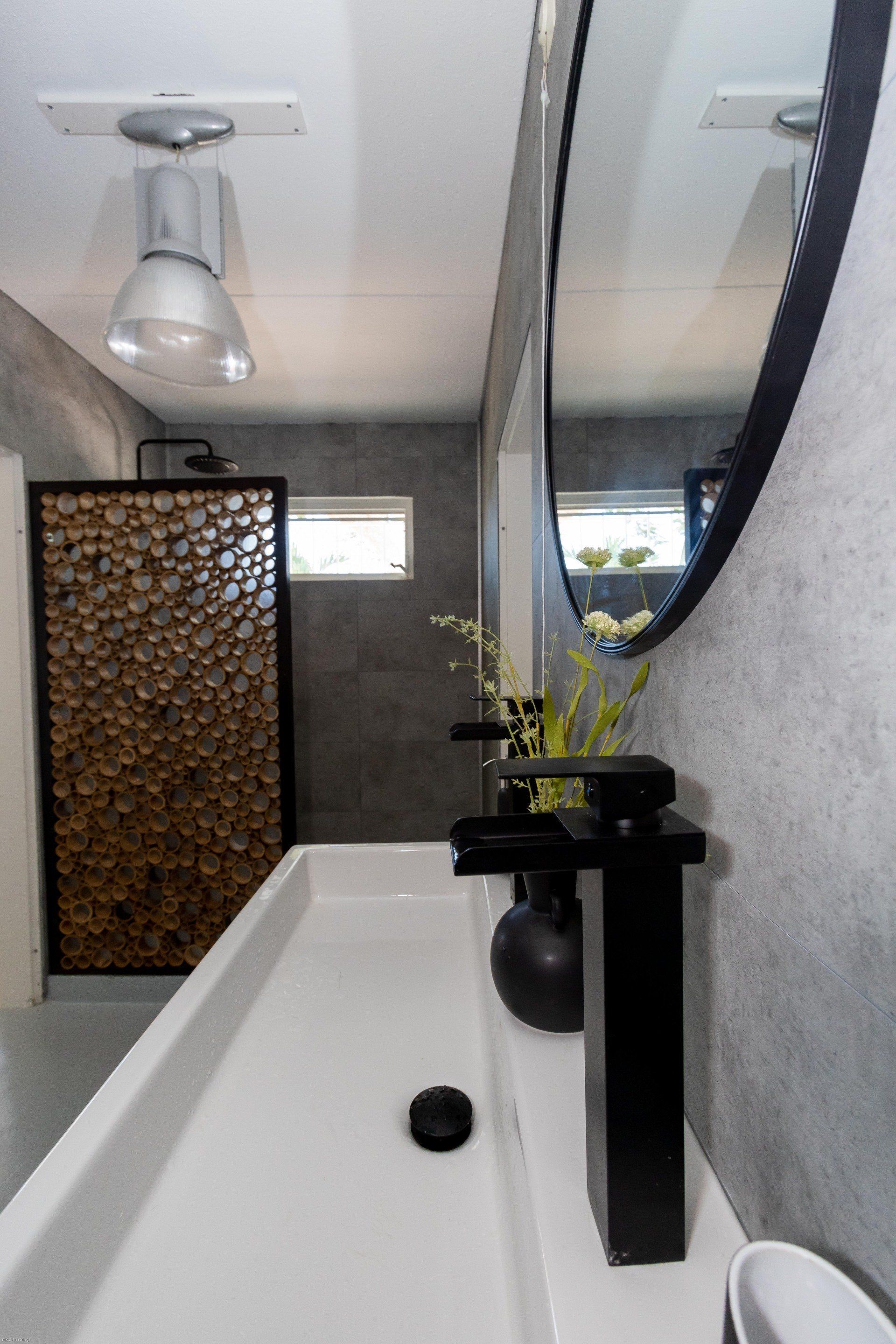 Moderne badkamer met lange witte wastafel, zwarte kraan, grote spiegel en douchewand met houten panelen.