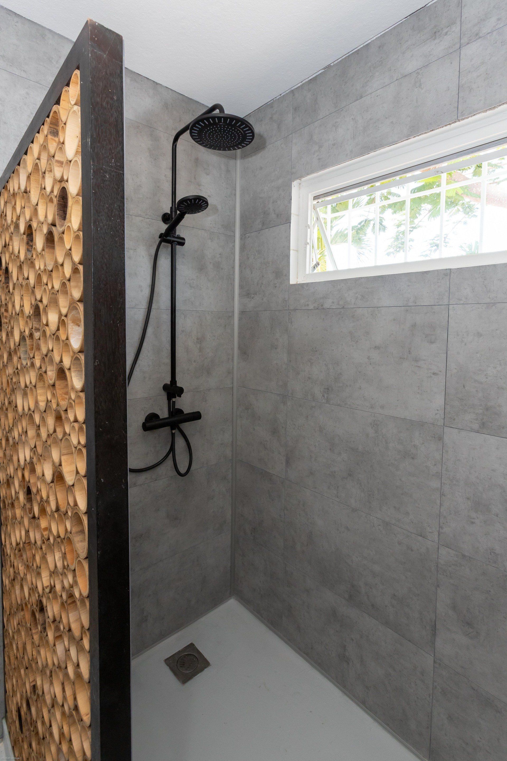Douche met zwarte kranen, grijze tegelwanden, een decoratief houten scherm en een klein raam.