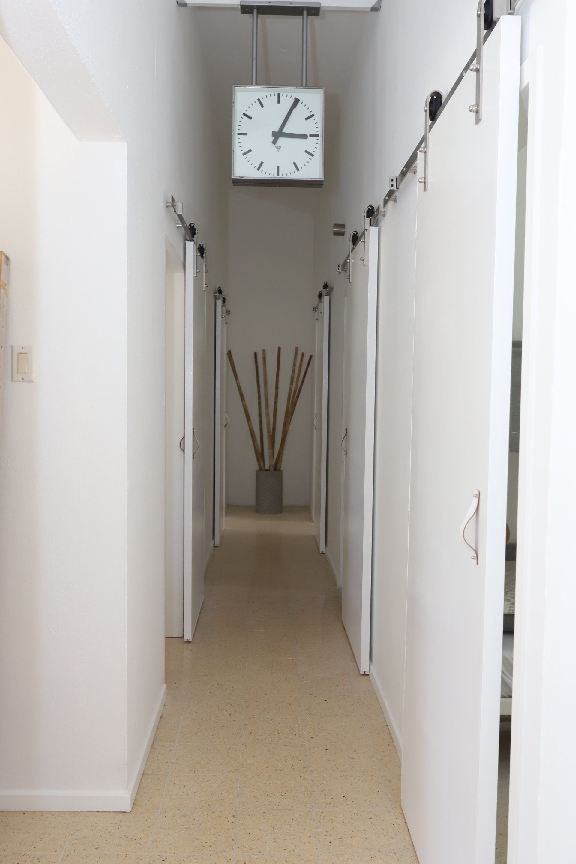 Smalle witte gang met deuren, een hangende klok en decoratieve stokken aan het einde.