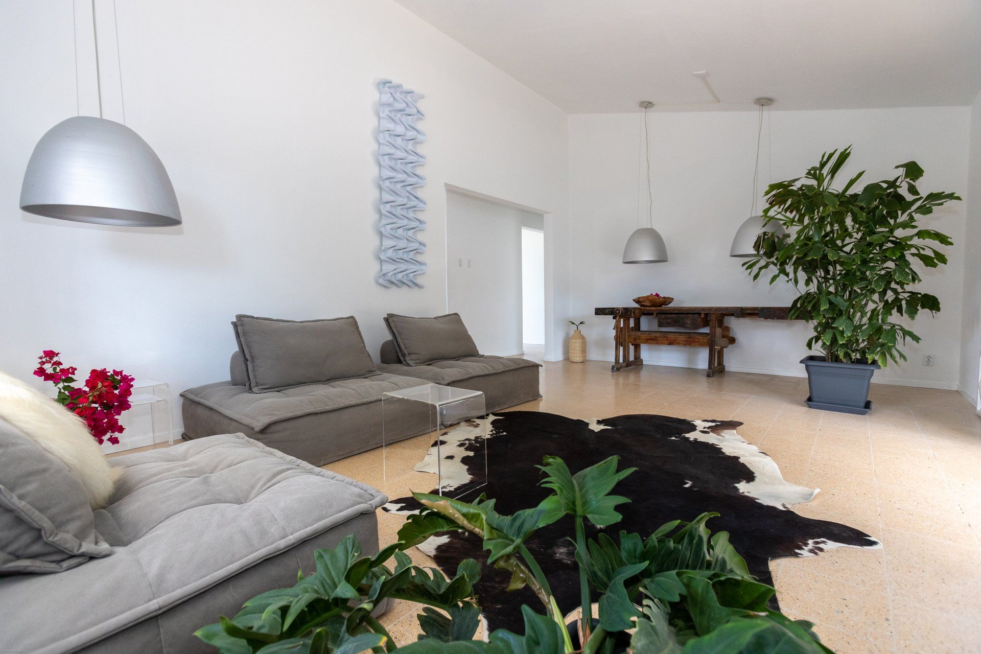 Ruime, lichte woonkamer met grijze modulaire banken, koeienhuid vloerkleed, grote plant en hangende zilverkleurige lampen.