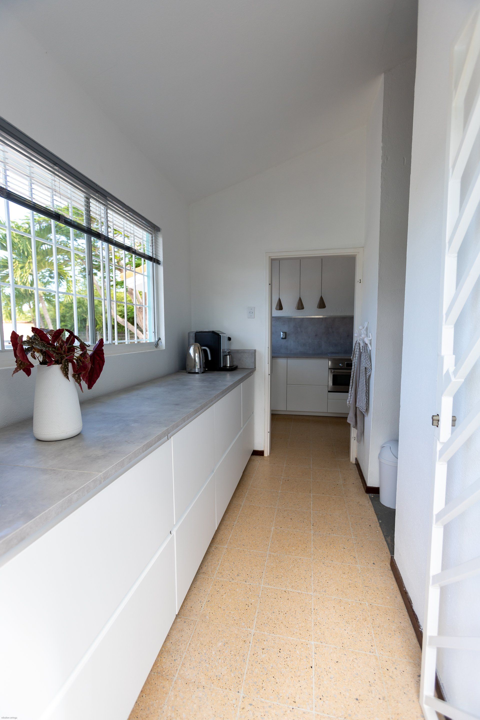 Lange, smalle, witte keuken met aanrechtblad, raam en deur naar een andere kamer. Betonnen vloer.