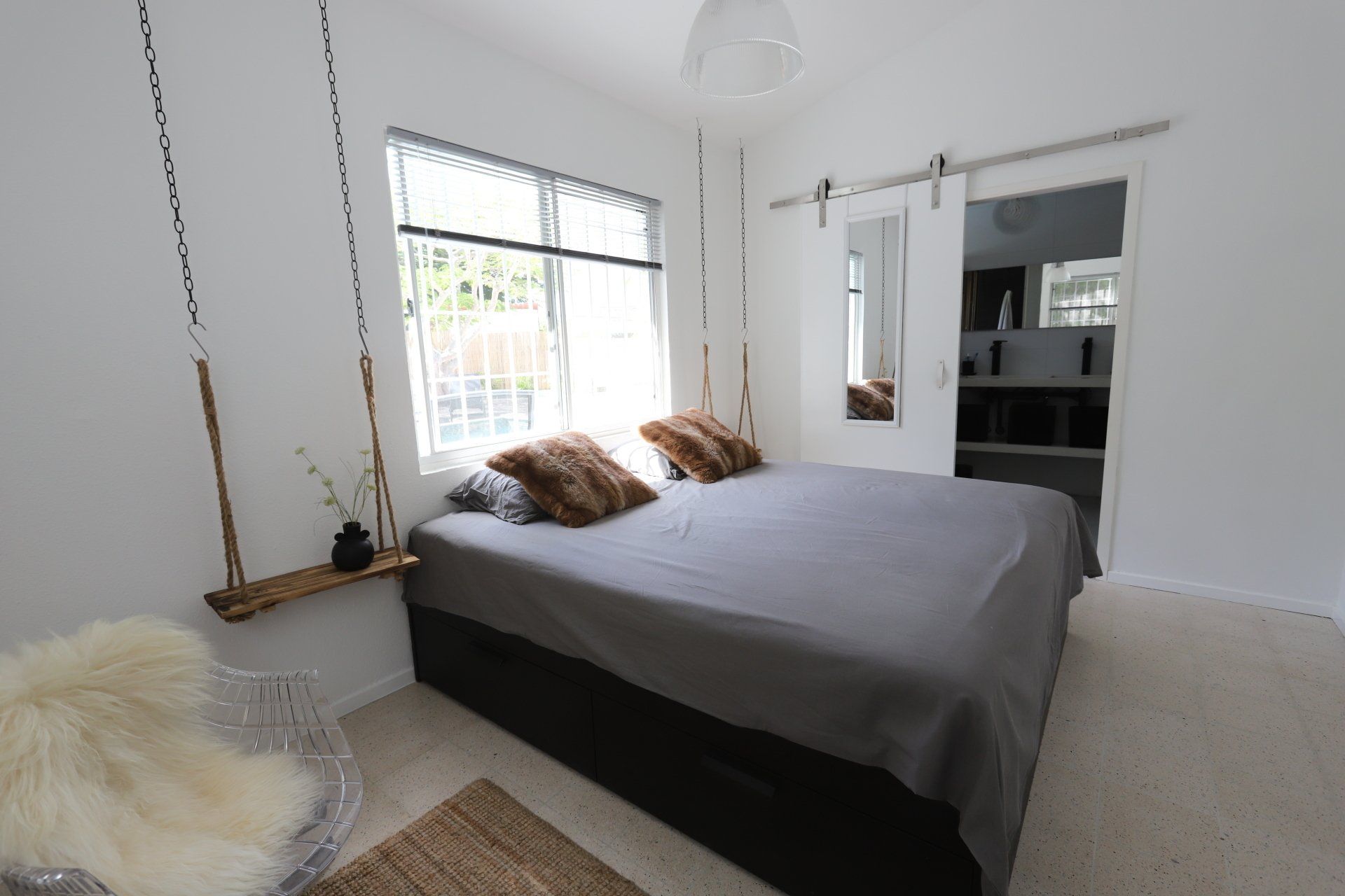 Slaapkamer met bed, schuifdeur, raam en schommel. Witte muren, grijs beddengoed, kussens van imitatiebont en een transparante stoel.