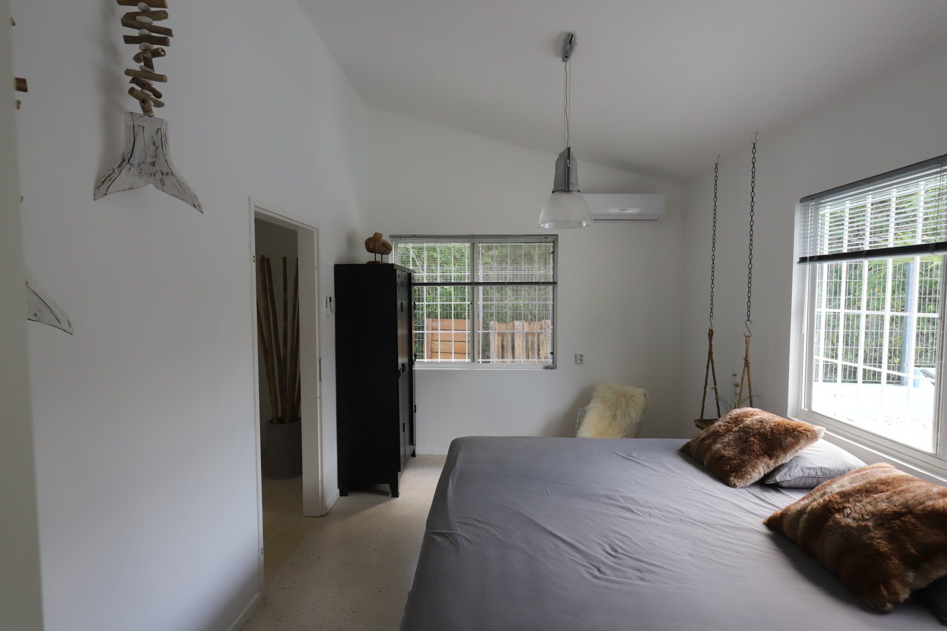 Slaapkamer met witte muren, een bed en decoratieve wanddecoratie. Een raam laat zonlicht binnen.