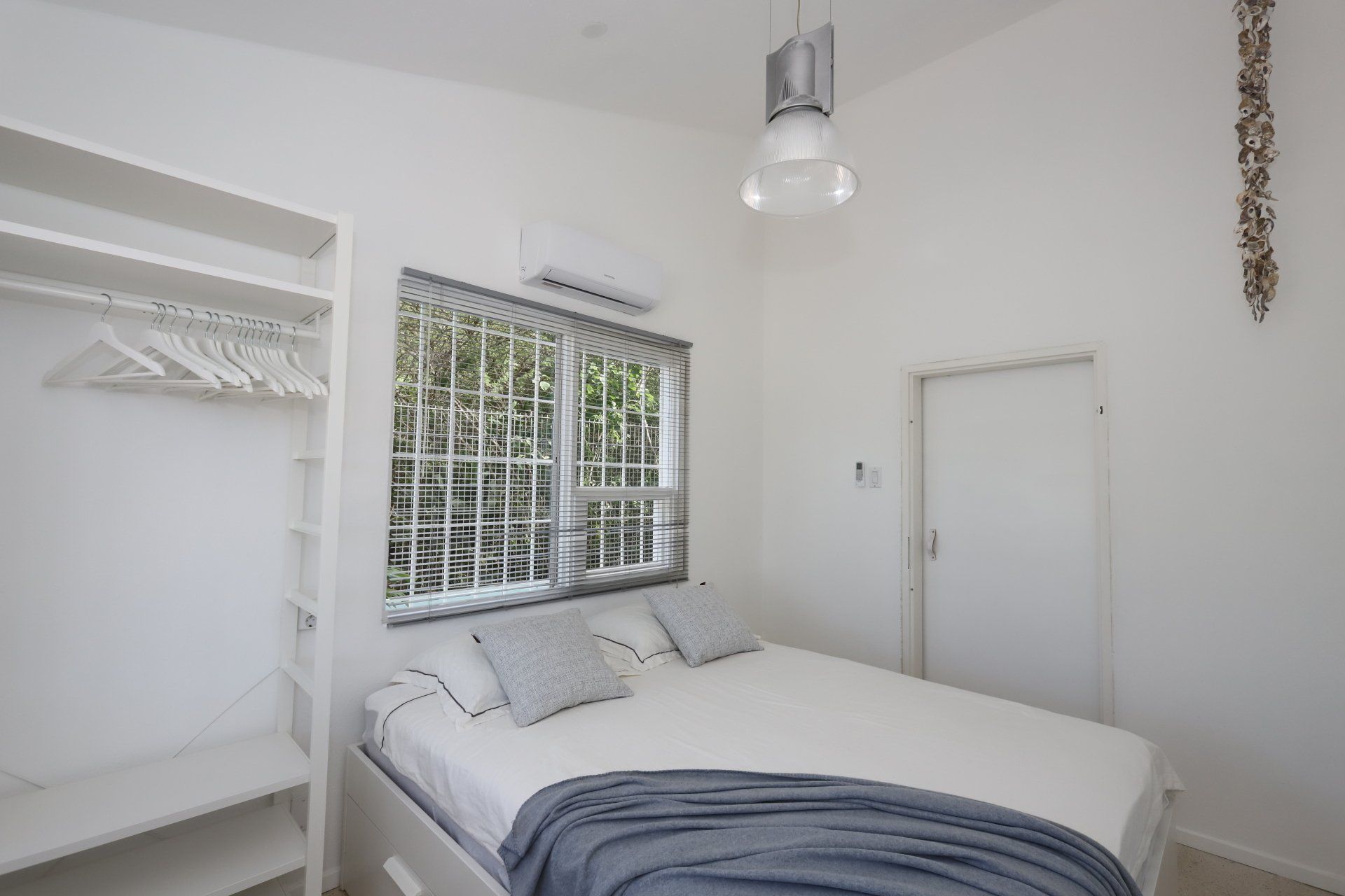 Slaapkamer met een bed, raam, open kast en een gesloten deur. Witte muren en meubels.