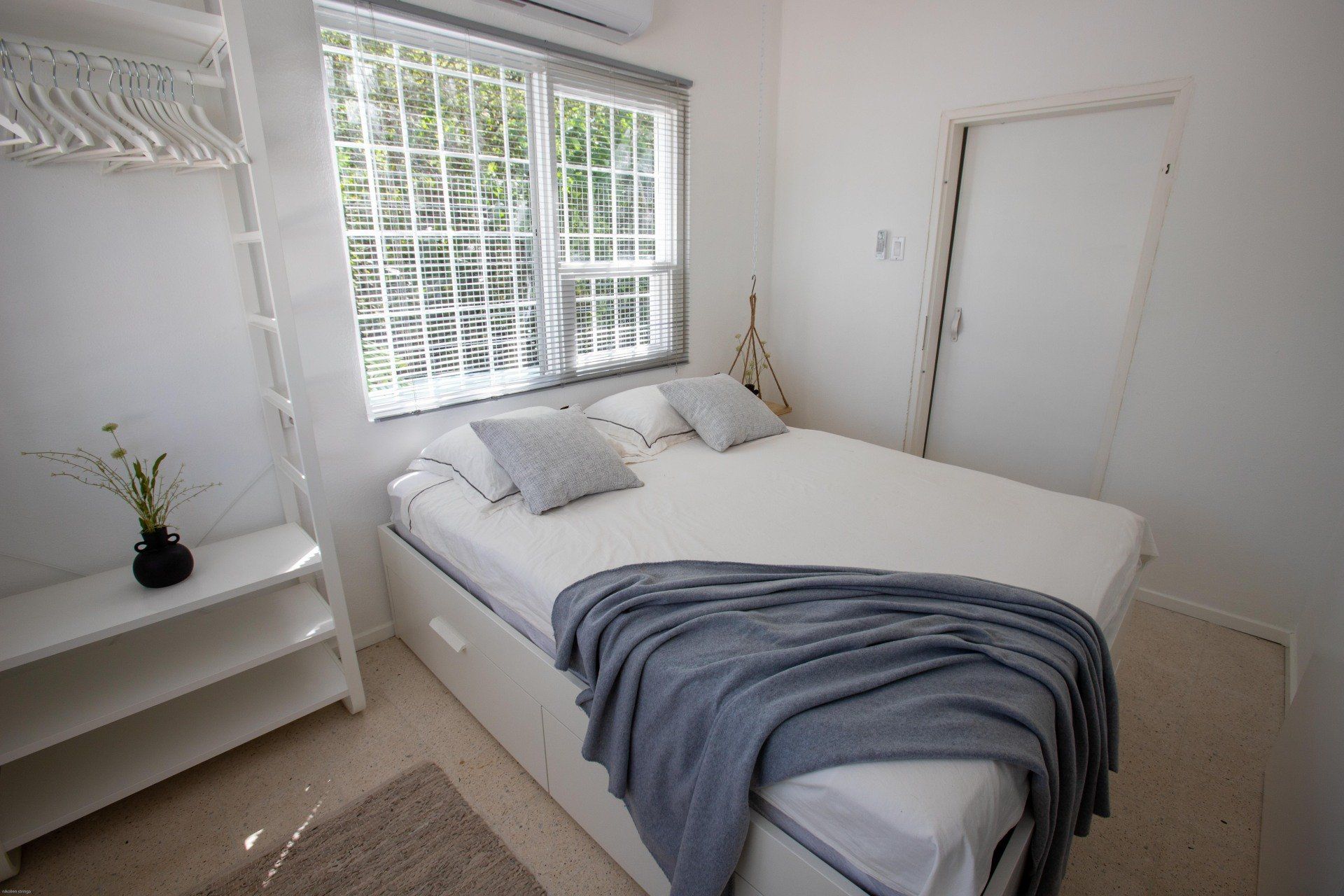 Witte slaapkamer met een bed, raam en kleine plankjes.