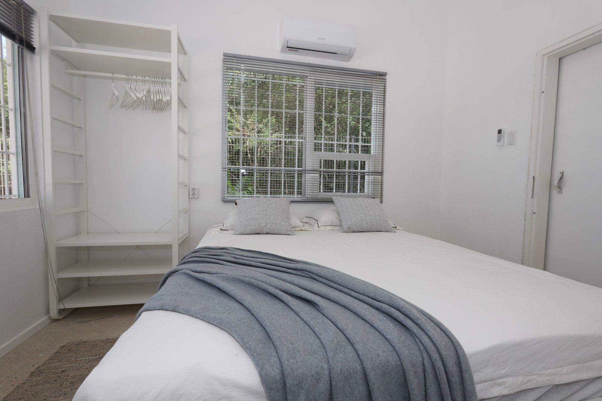 Witte slaapkamer met bed, open kledingkast en raam met uitzicht op bomen.