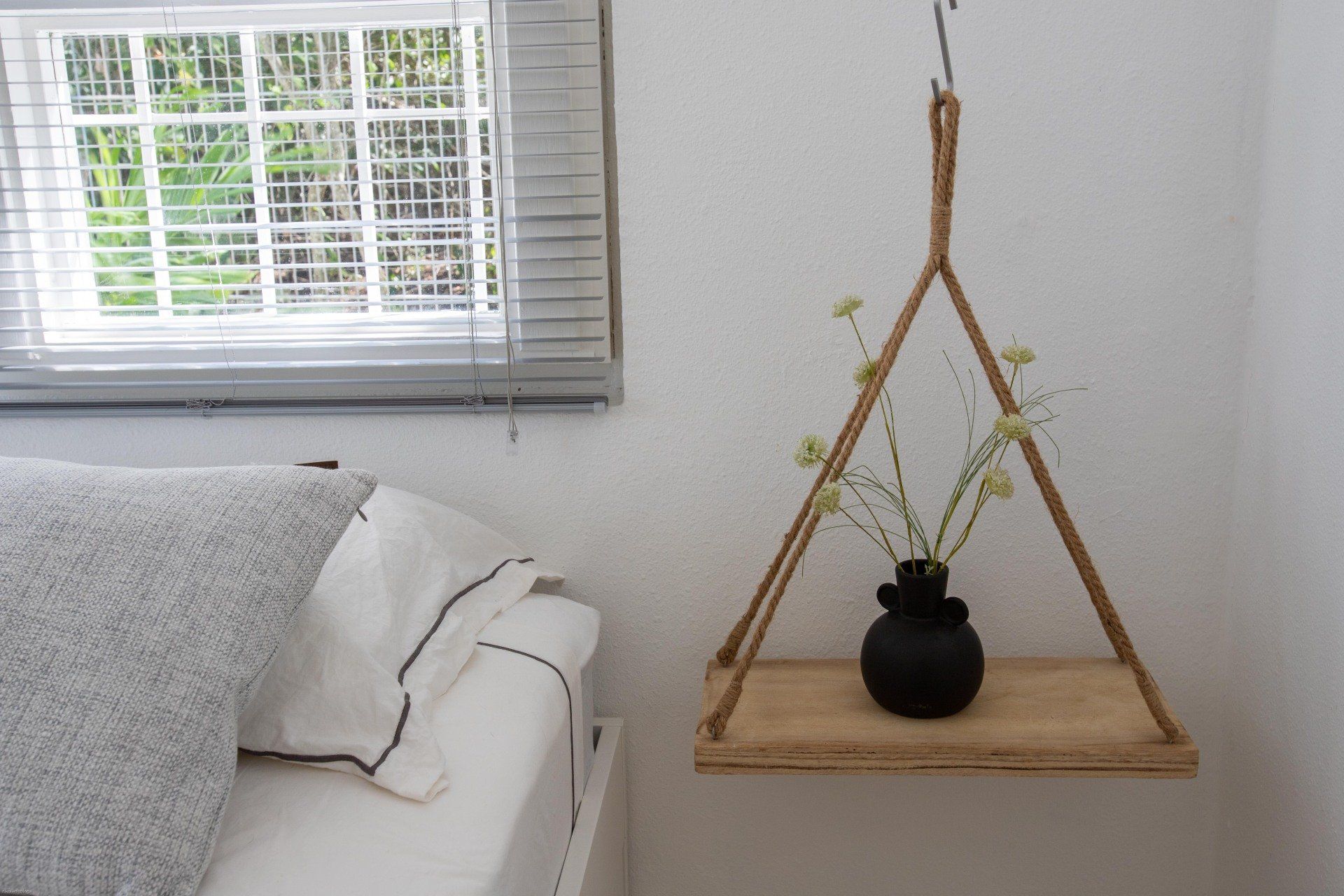 Hangende houten plank met zwarte vaas en bloemen. Touw ondersteunt de plank, vlakbij een bed en een raam met jaloezieën.