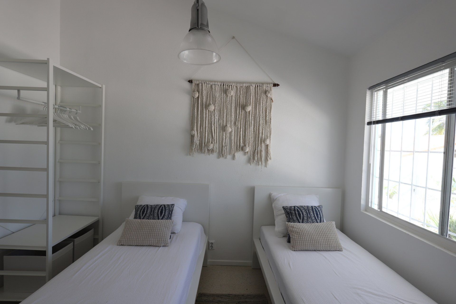 Twee eenpersoonsbedden met wit beddengoed in een kamer, een macramé wandkleed en een raam.