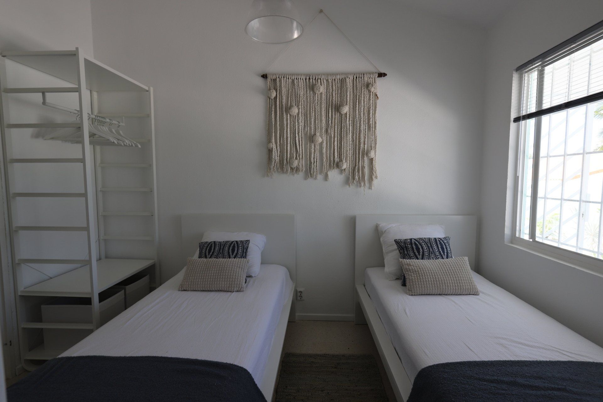 Twee eenpersoonsbedden in een witte slaapkamer met een macramé wandkleed en een raam met jaloezieën.