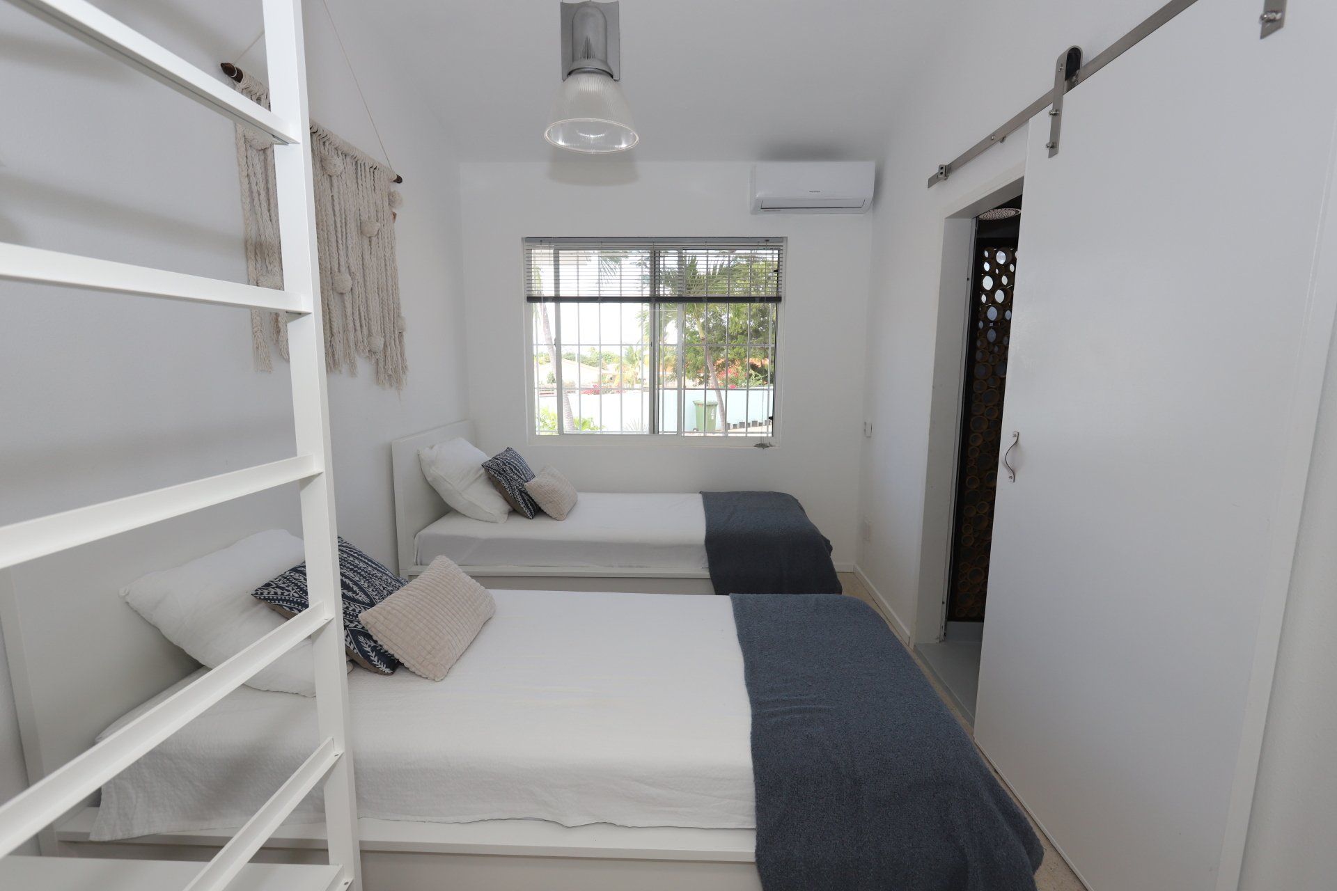 Witte slaapkamer met twee eenpersoonsbedden, een klein raam, een schuifdeur en een macramé wandkleed.