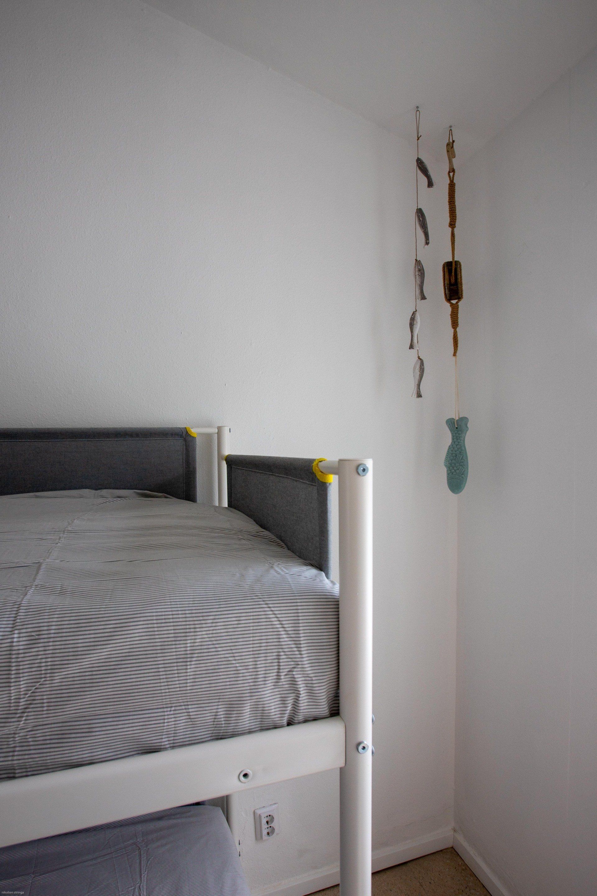 Een wit stapelbed met grijs beddengoed in een hoek van een witte kamer, met daar vlakbij een decoratieve mobiel.