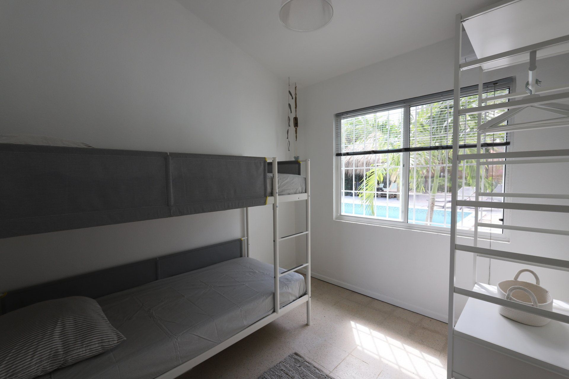 Stapelbedden in een witte slaapkamer met een raam dat uitkijkt op een zwembad en planken aan de rechterkant.