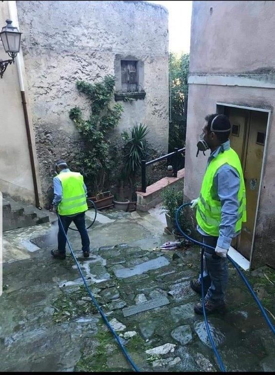 Due operai con i gilet lavano a pressione un vicolo in pietra con vecchi edifici.