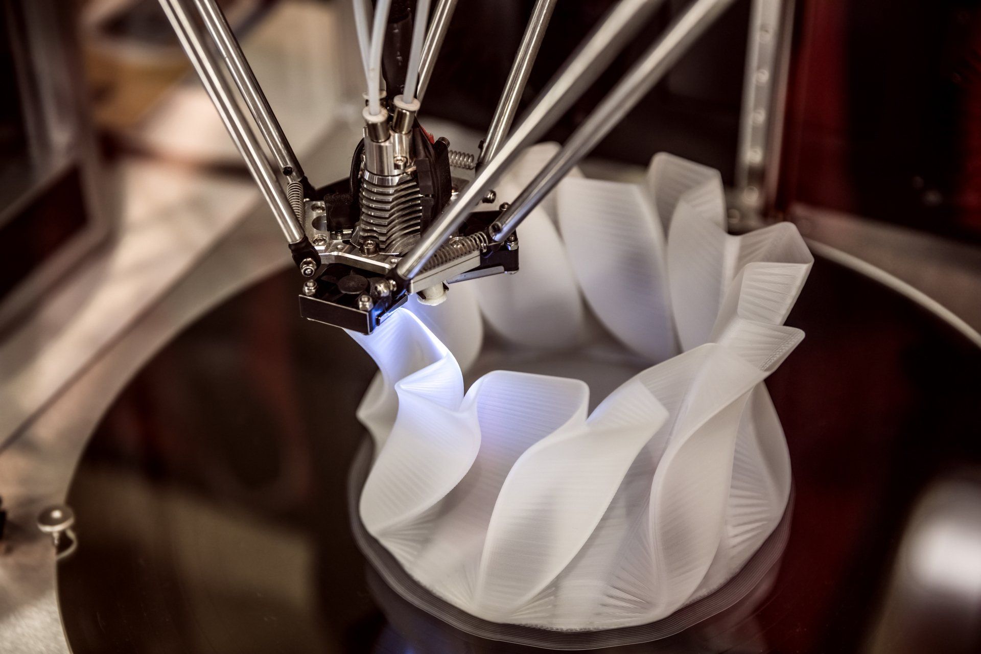 A 3D printer head depositing white filament to build a tall, wavy-textured cylindrical structure on a dark circular base.
