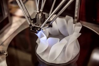 A 3D printer head depositing white plastic to create a wavy, translucent cylindrical structure on a dark circular platform.