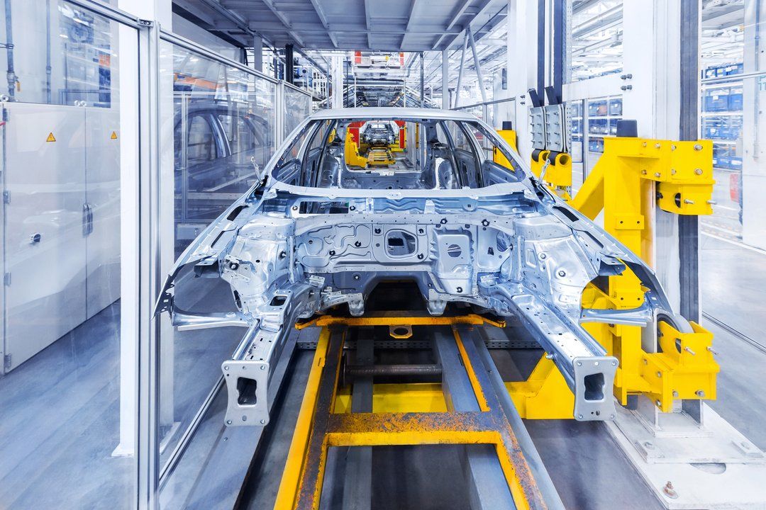 A metallic car body frame moves along a bright yellow industrial assembly line in a factory.