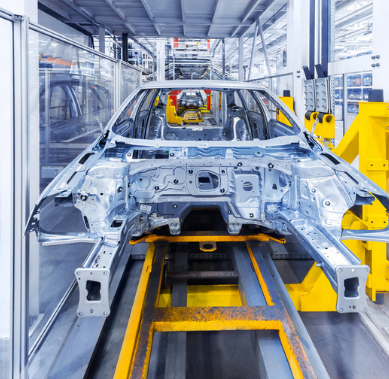 A metallic car body frame moves along a bright yellow assembly line in a modern manufacturing factory.