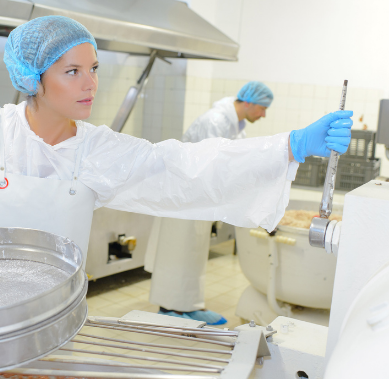 Two people in white protective gear and blue hairnets operate industrial food processing equipment in a sterile kitchen.